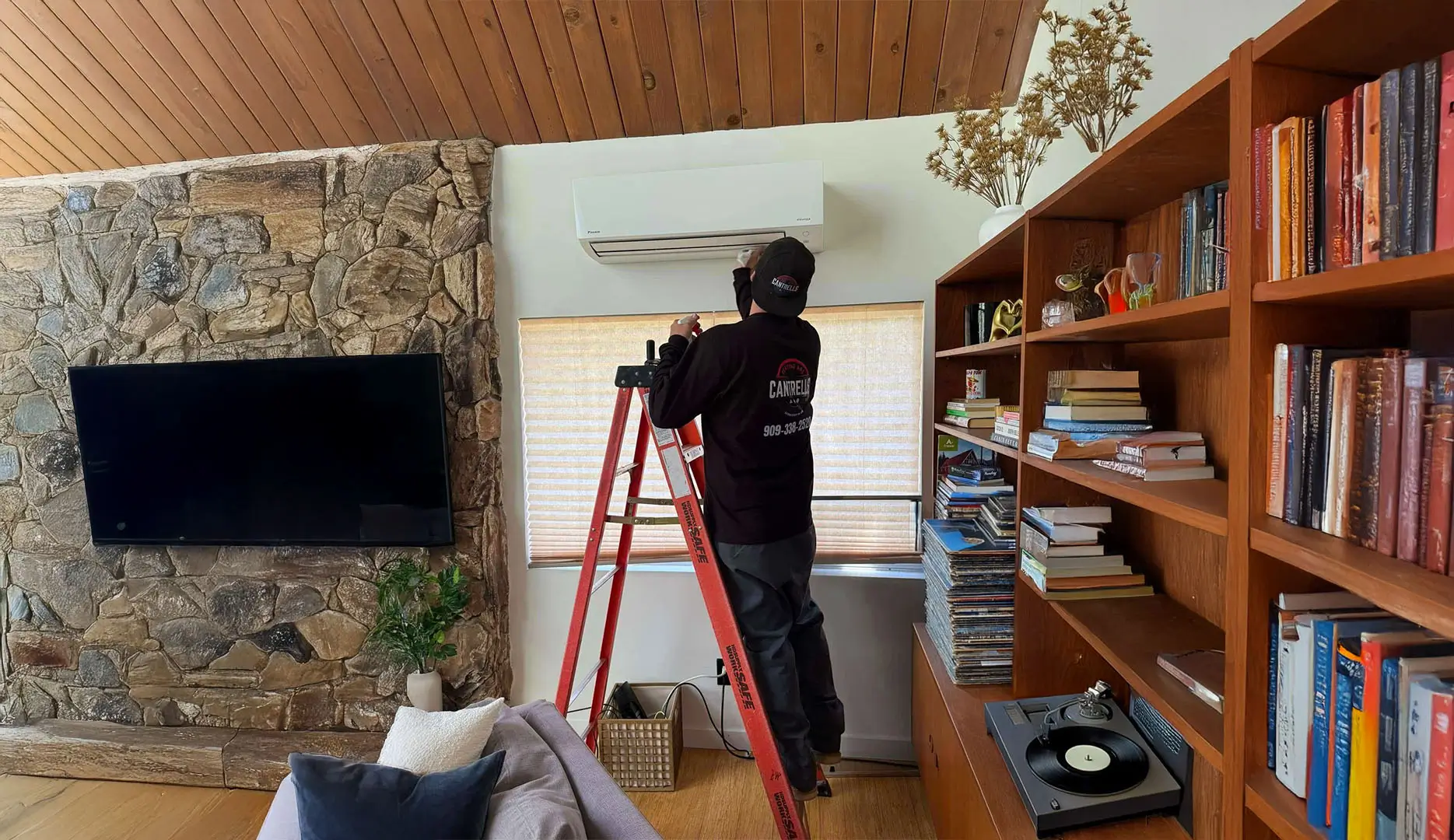 AC expert installing a new air conditioning unit on the wall near the ceiling and also see a big screen flat TV and a book shelf filled with books.