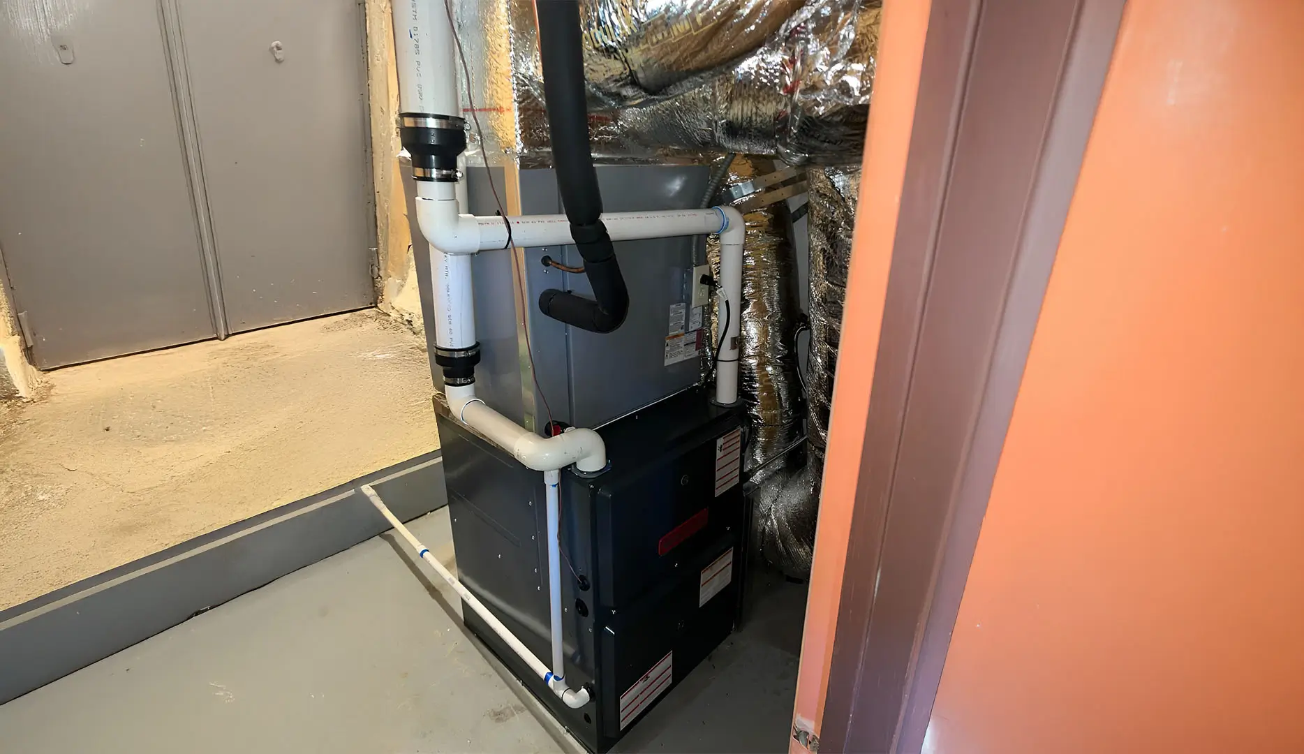 Heater in a basement with a red door and wall to the right of it and two metal gray doors to the left of it.