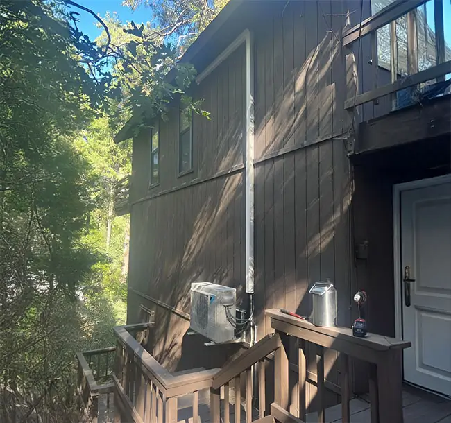 Ductless air conditioning system set up outside a two story home surrounded by trees.