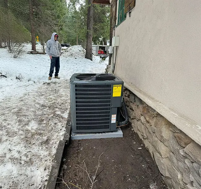 A young male contractor looks at a lone AC unit outside a home with snow on the ground.