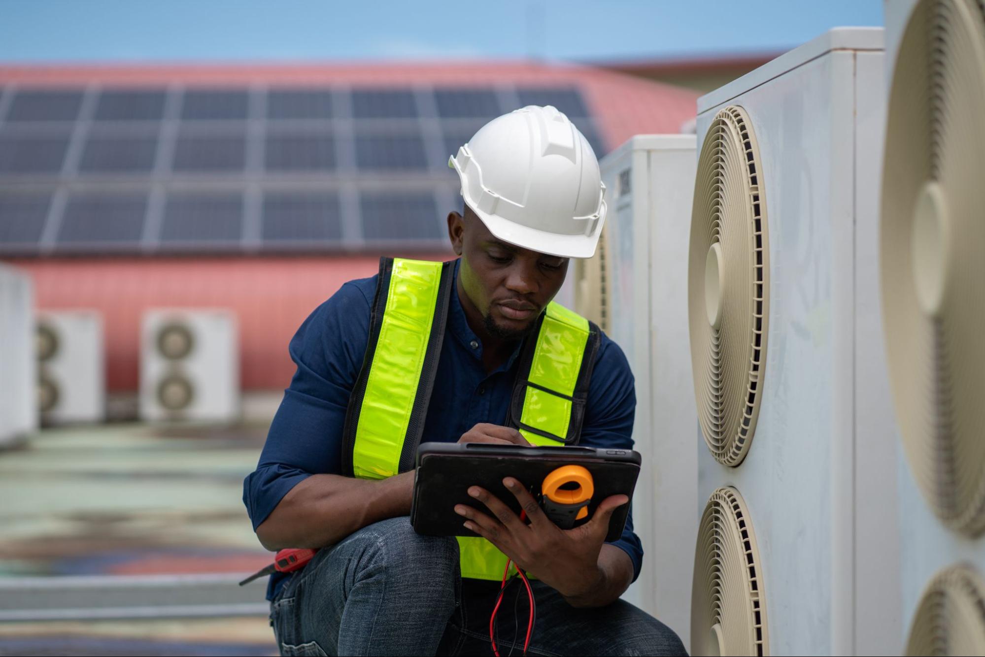 A Black HVAC technician in a yellow hard hat inspects the commercial air conditioning unit outside a building
