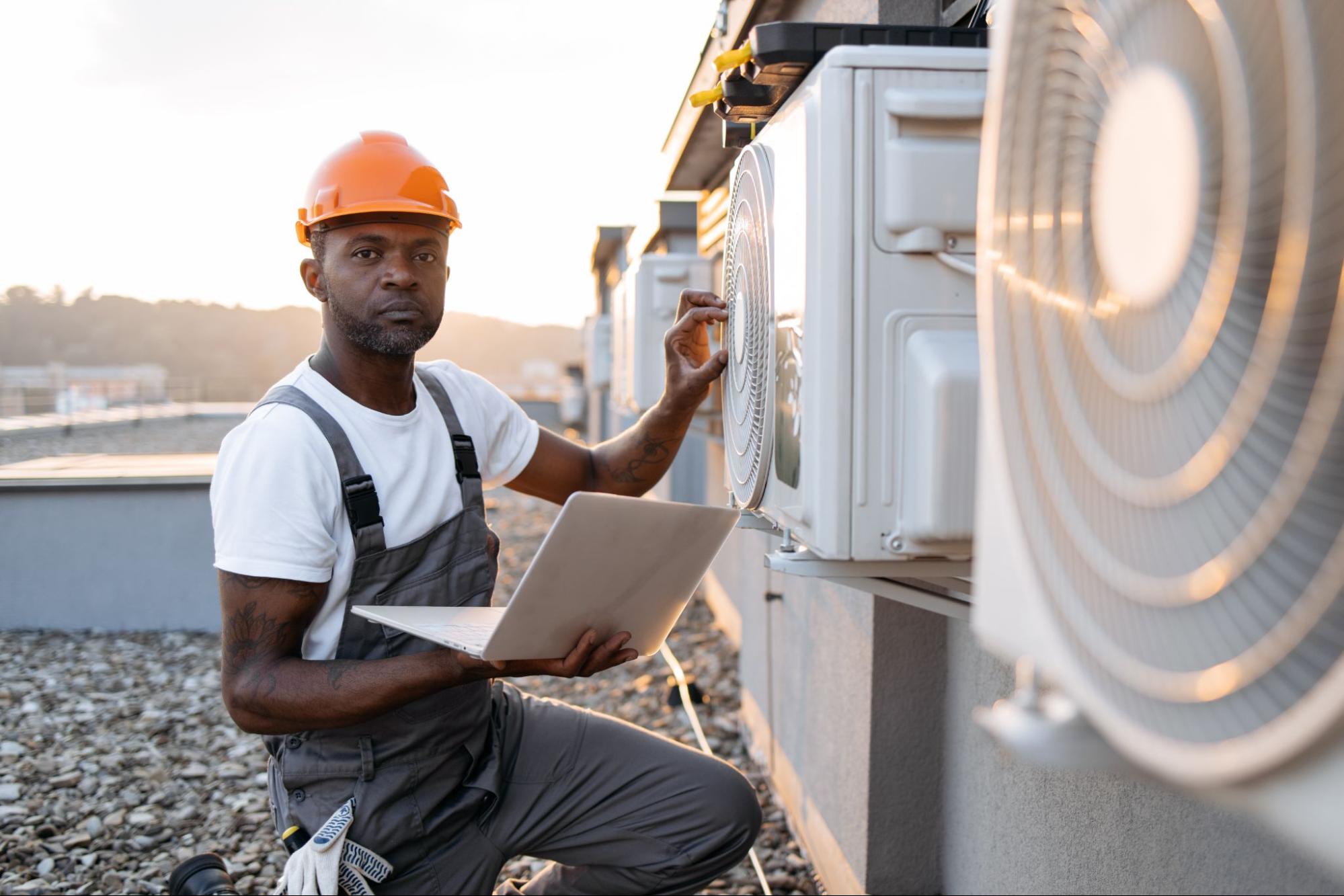A man holds a laptop while repairing a rooftop AC unit.