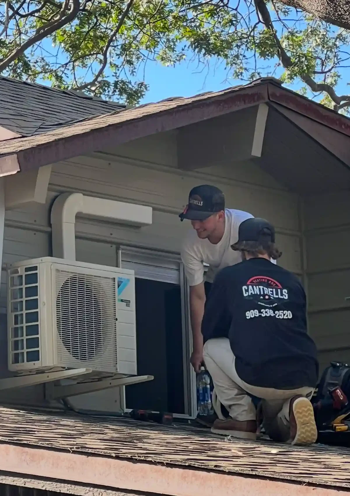 Two male HVAC contractors on top of a room install a single mini split air conditioning unit.